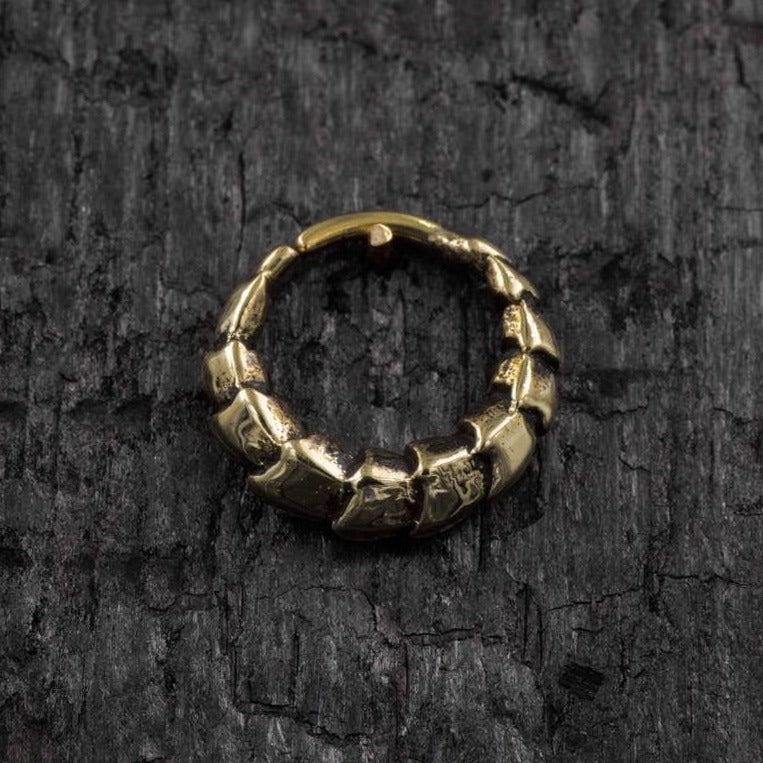 The AARO Cyborg Seamless Hoop Septum Nose Ring in Gold by Kaya Movement, featuring an 18 gauge design and an irregular, segmented appearance, rests on a textured, dark gray surface. The rough, uneven segments give the ring a rugged, ancient appearance.